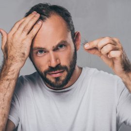 bearded middle aged man holding fallen hair and looking at camera isolated on grey